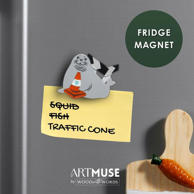 A refrigerator magnet inspired by Neil the Seal holding an orange traffic cone, attached to a yellow sticky note on a stainless steel fridge. A dark green circle reads 'FRIDGE MAGNET,' and the ArtMuse by Wood & Words logo appears at the bottom.