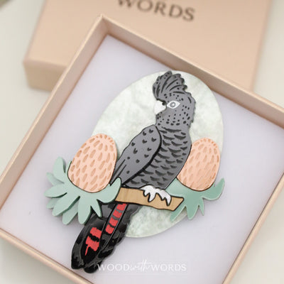 A brooch depicting a Red-tailed Black Cockatoo perched on a branch, presented in a gift box.